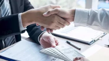 Dos personas estrechando manos sobre un escritorio con documentos y dinero.