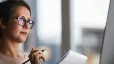 Mujer con gafas escribe en un cuaderno frente a una computadora.