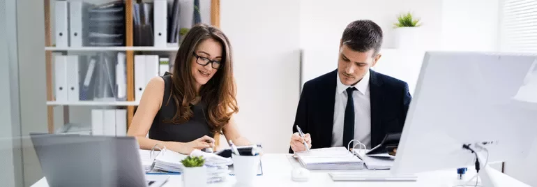Dos personas trabajando en una oficina con documentos y computadoras.