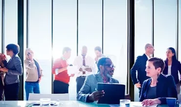 Personas en reunión de negocios, conversando junto a ventanas grandes.