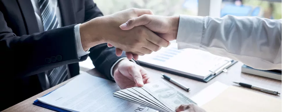 Dos personas estrechando manos sobre un escritorio con documentos y dinero.