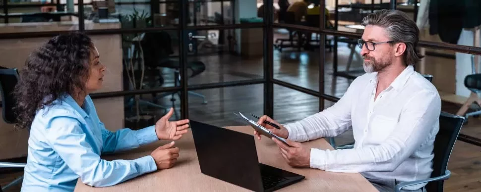 Dos personas conversando en una oficina con portátil sobre la mesa.