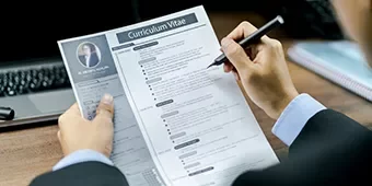 Persona revisando un currículum en su escritorio, con un ordenador portátil al fondo.